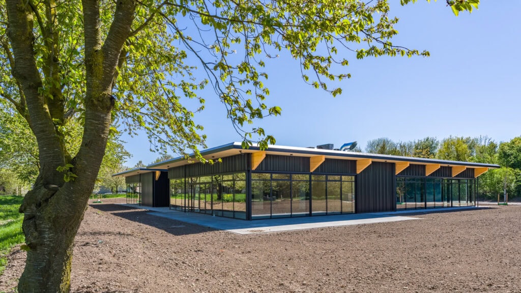 Moderne sporthal met grote glazen ramen en houten accenten, omgeven door groen en een open veld.