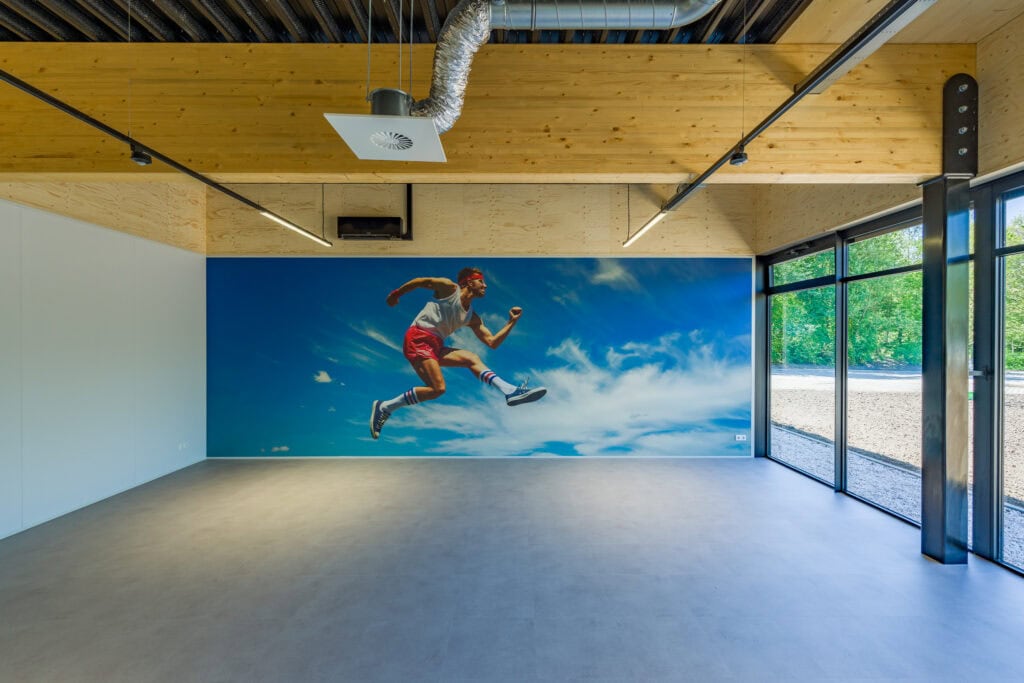 Ruime sportzaal met grote muurschildering van een springende persoon en grote ramen.