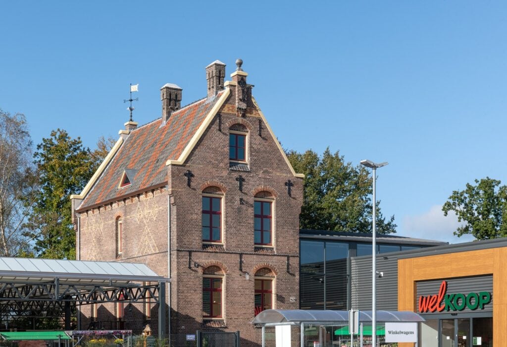 Een oud bakstenen gebouw met karakter, naast een moderne supermarkt, onder een heldere blauwe hemel.