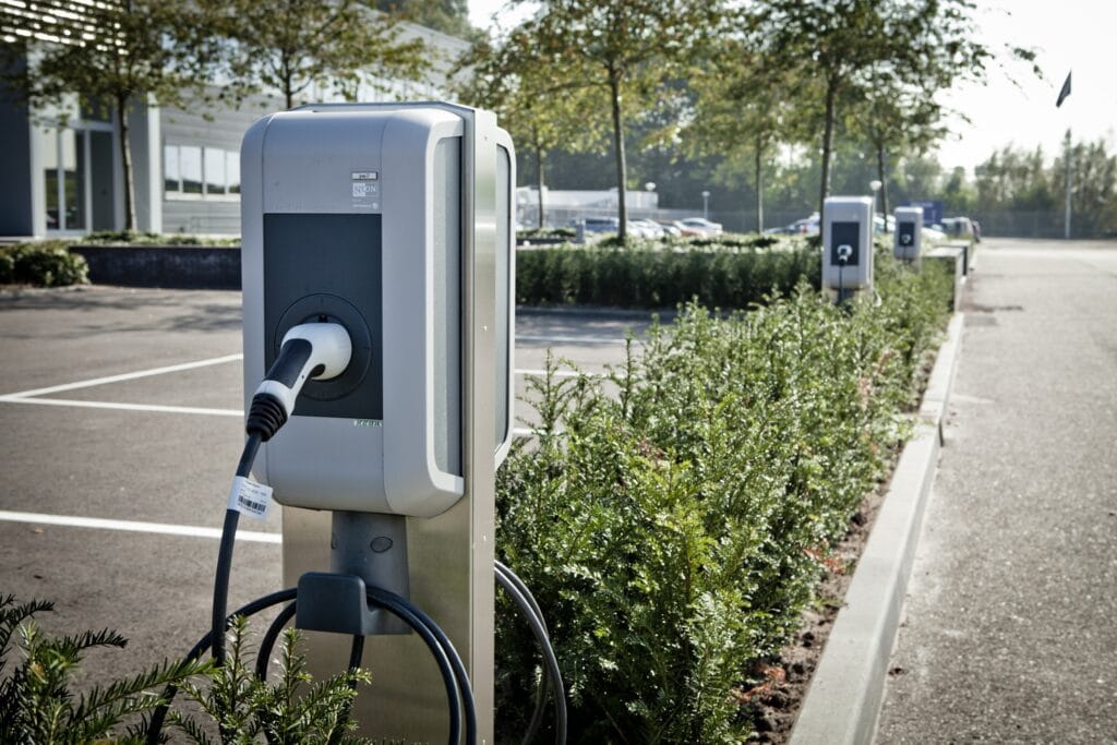 Elektrische laadpaal in een parkeerplaats met groene struiken.