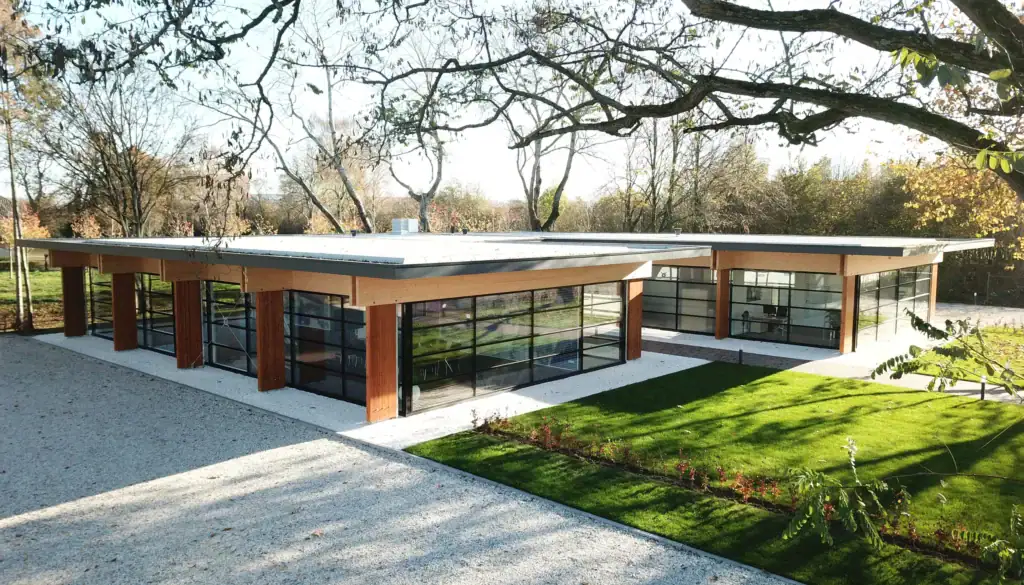 Moderne bouw met grote glazen ramen en plat dak, omgeven door groen landschap.
